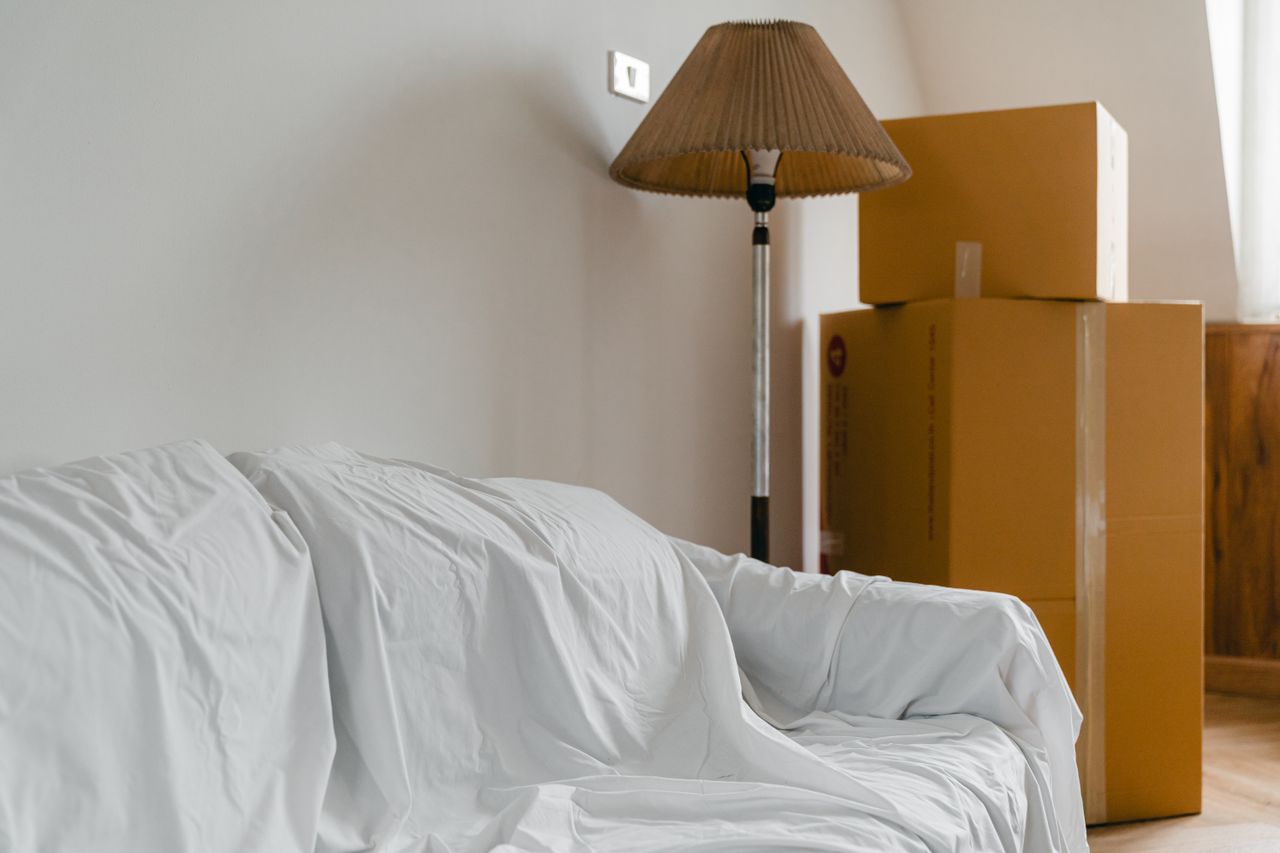 Services-03 Covered with sheet sofa and floor lamp and packed cardboard boxes in light spacious room of new contemporary house